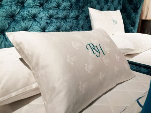 Kopfkissen mit edler Stickerei „RH“ vor einem blau gepolsterten Betthaupt